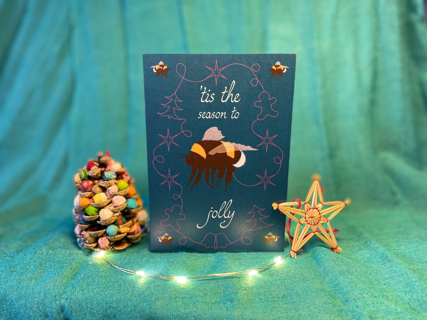 A photo of a Christmas card against a teal backdrop with a Christmas decoration either side. On the card itself is a flying bumblebee in the centre with a miniature bumblebees in each corner. The centre bumblebee's flight trail loops around it in the shape of stars, gingerbread men and Christmas trees. Above the centre bee it says "'Tis the season to" and beneath it, it says "jolly". The card's background is deep turquoise.