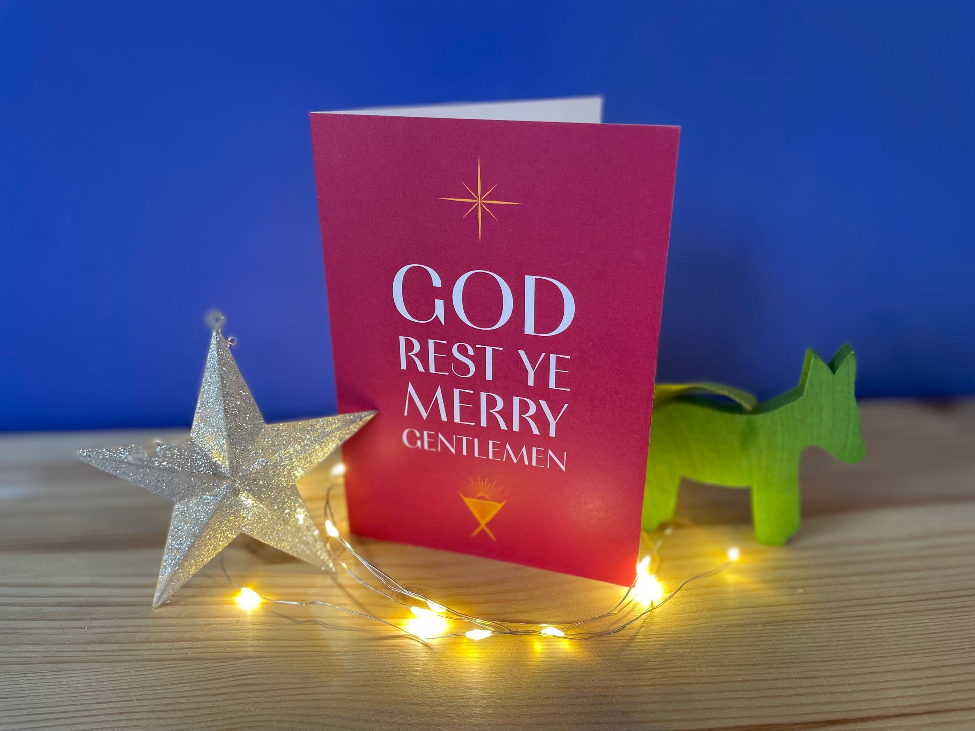 A photo of a Christmas card which sits on a wooden shelf against a deep purple-blue wall. There is one Christmas decoration either side of it and lights around it. The card is A6 portrait with a crimson background. In white writing it says "God rest ye merry gentlemen". There is a golden manger beneath the text and a golden star above the text.