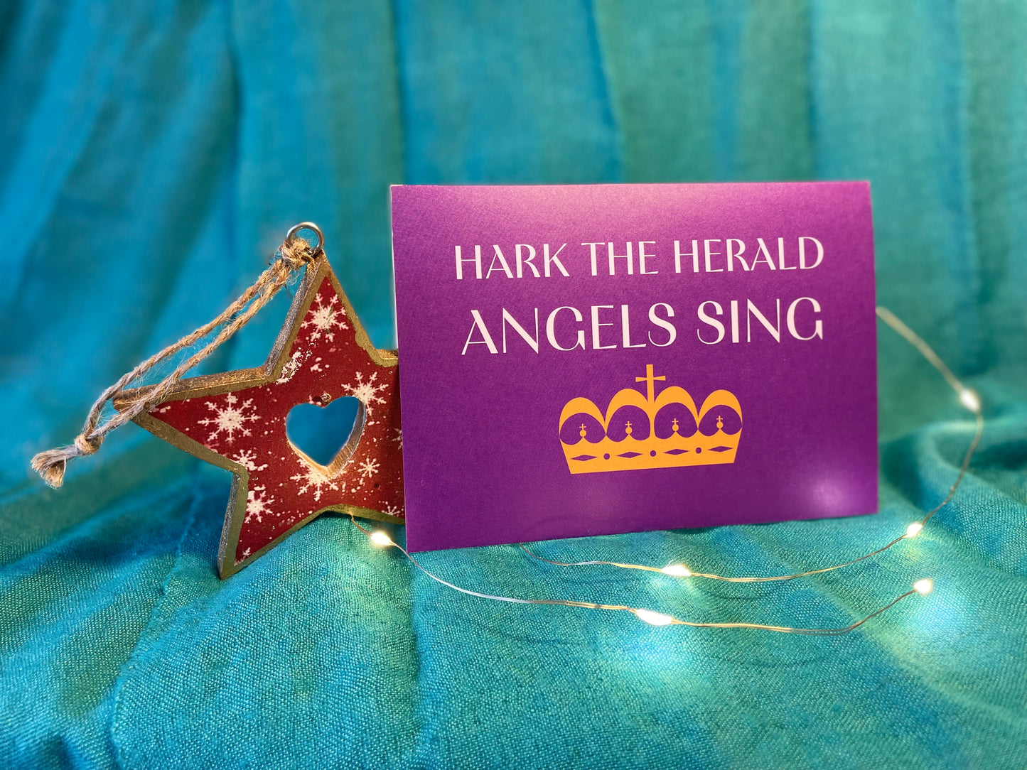 A photo of a Christmas card which sits on a teal backdrop with a Christmas decoration and lights around it. The card is A6 landscape with a deep purple background. In white writing it says "Hark the herald angels sing" and there is a golden crown beneath the text.