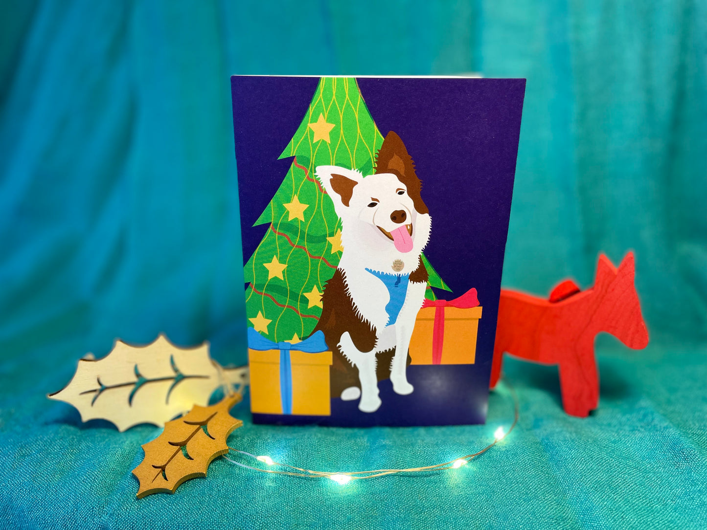 A Christmas card sits between some Christmas decorations and lights on a teal background. The card is A6 portrait and show a red-and-white Border Collie sits between two presents and in front of a decorated Christmas tree. She's wearing a harness which matches one of the present's ribbons. Her head is cocked and she appears to be smiling at you!