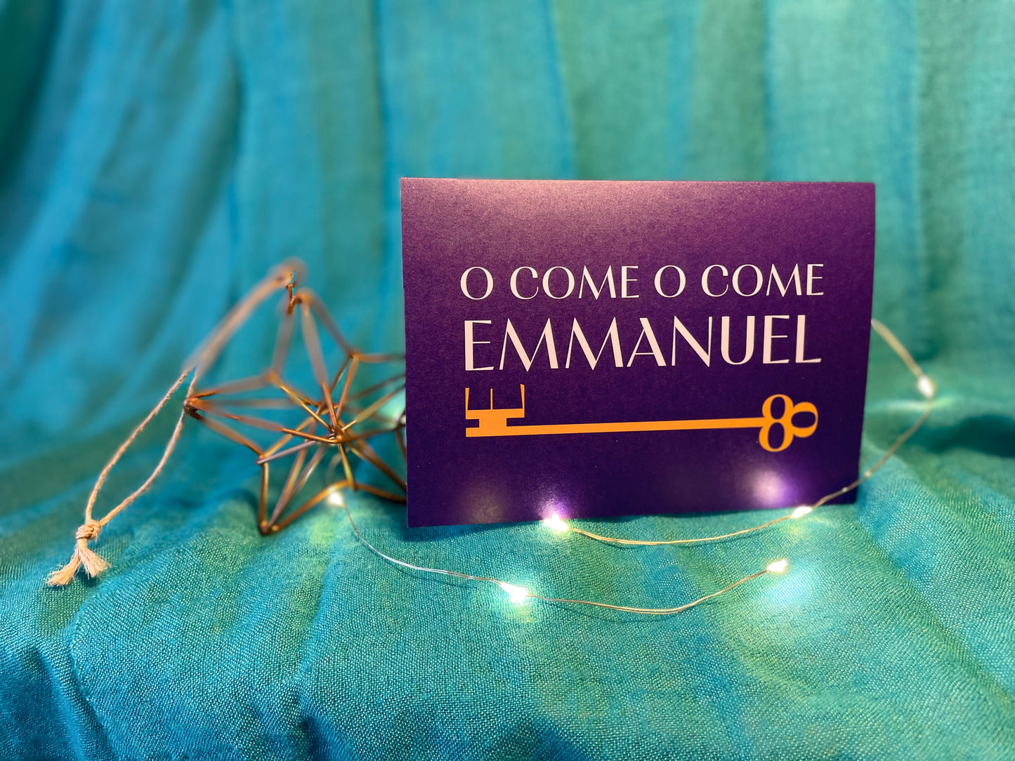 A photo of a Christmas card which sits on a teal backdrop with a Christmas decoration beside it and lights around it. The card is A6 landscape with a deep purple background. In white writing it says “O come O come Emmanuel” and there is a golden key beneath the text.